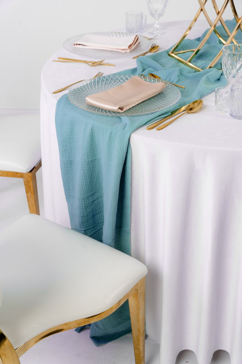 Długi bieżnik muślinowy w kolorze baby blue, miękko udrapowany na drewnianym stole weselnym – lekka, półtransparentna tkanina w stylu Coastal Chic, wynajem tekstyliów eventowych