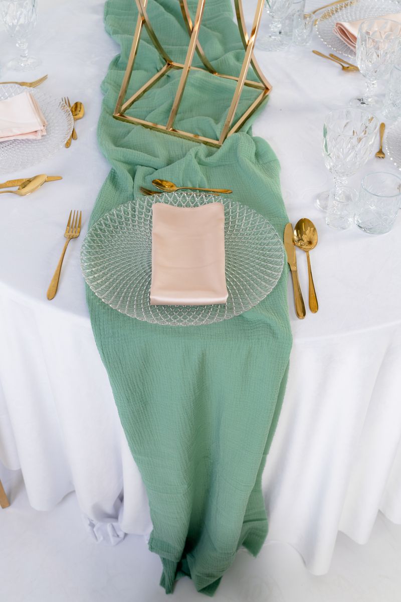 Szałwiowy bieżnik muślinowy ułożony w marszczenia na surowym drewnianym stole – dekoracja weselna w stylu boho i greenery na wynajem