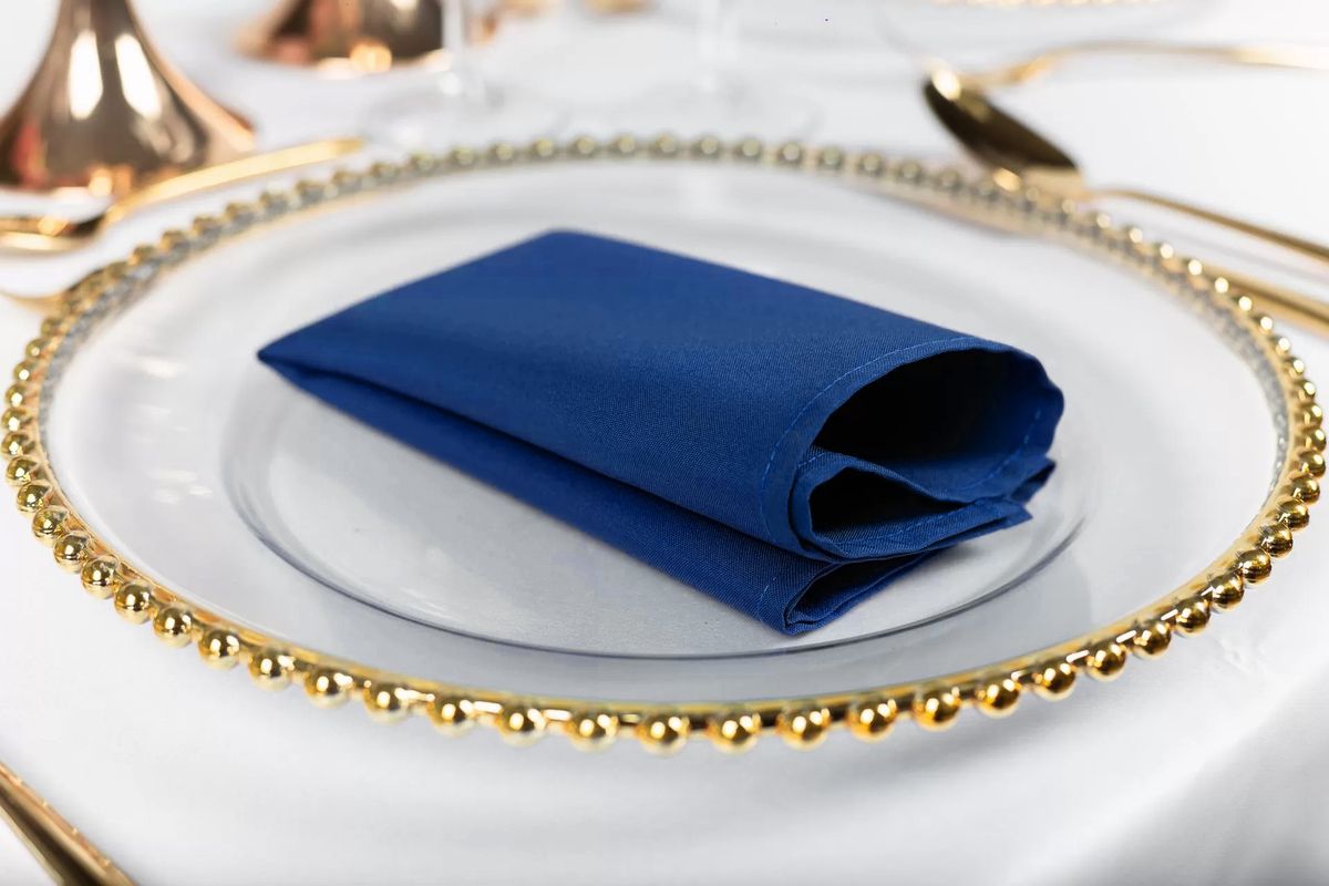 Chabrowa serwetka poliestrowa złożona w ostrosłup na białym talerzu ze złotym rantem – dekoracja stołu w kolorze royal blue, wynajem tekstyliów