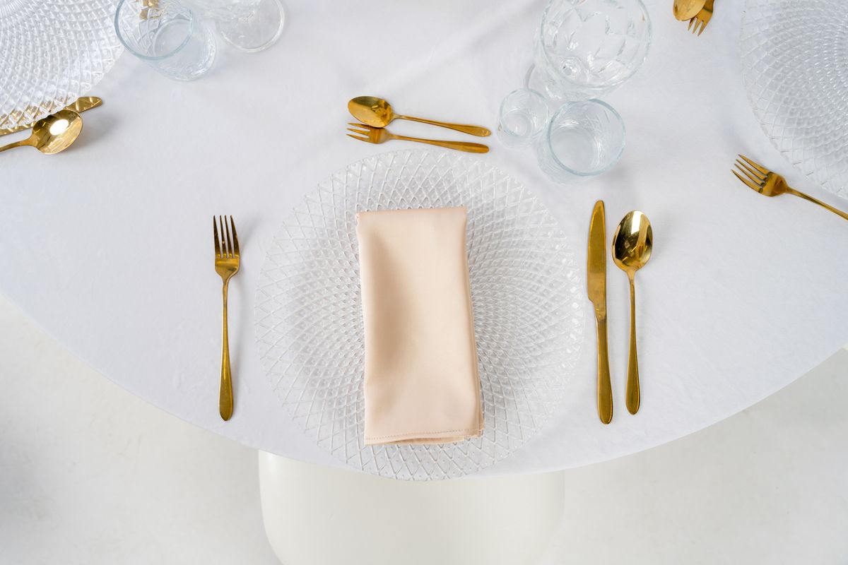 Beżowe serwetki satynowe na wynajem, ułożone na białym obrusie ze złotą zastawą i szampańskimi kieliszkami – luksusowa dekoracja stołu w stylu Quiet Luxury