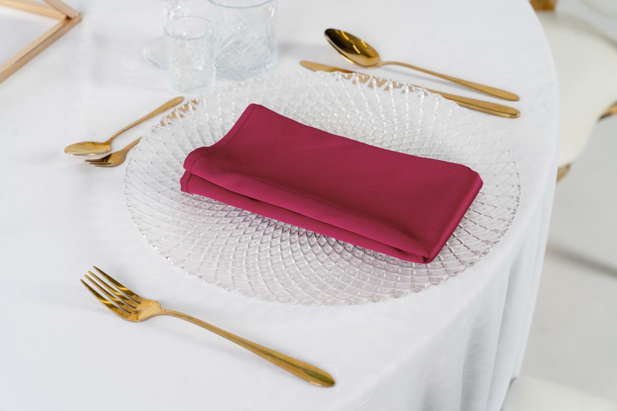 Bordowe serwetki satynowe na wynajem, ułożone na złotych podtalerzach – elegancka dekoracja stołu weselnego w stylu Burgundy Glamour.
