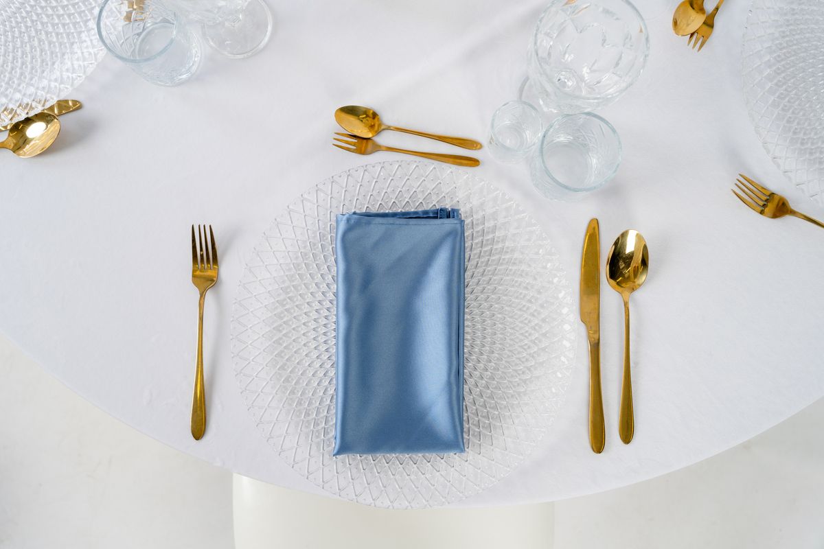 Satynowe serwetki dusty blue na wynajem, ułożone na białym obrusie ze srebrnymi sztućcami – elegancka dekoracja stołu weselnego w stylu Fine Art