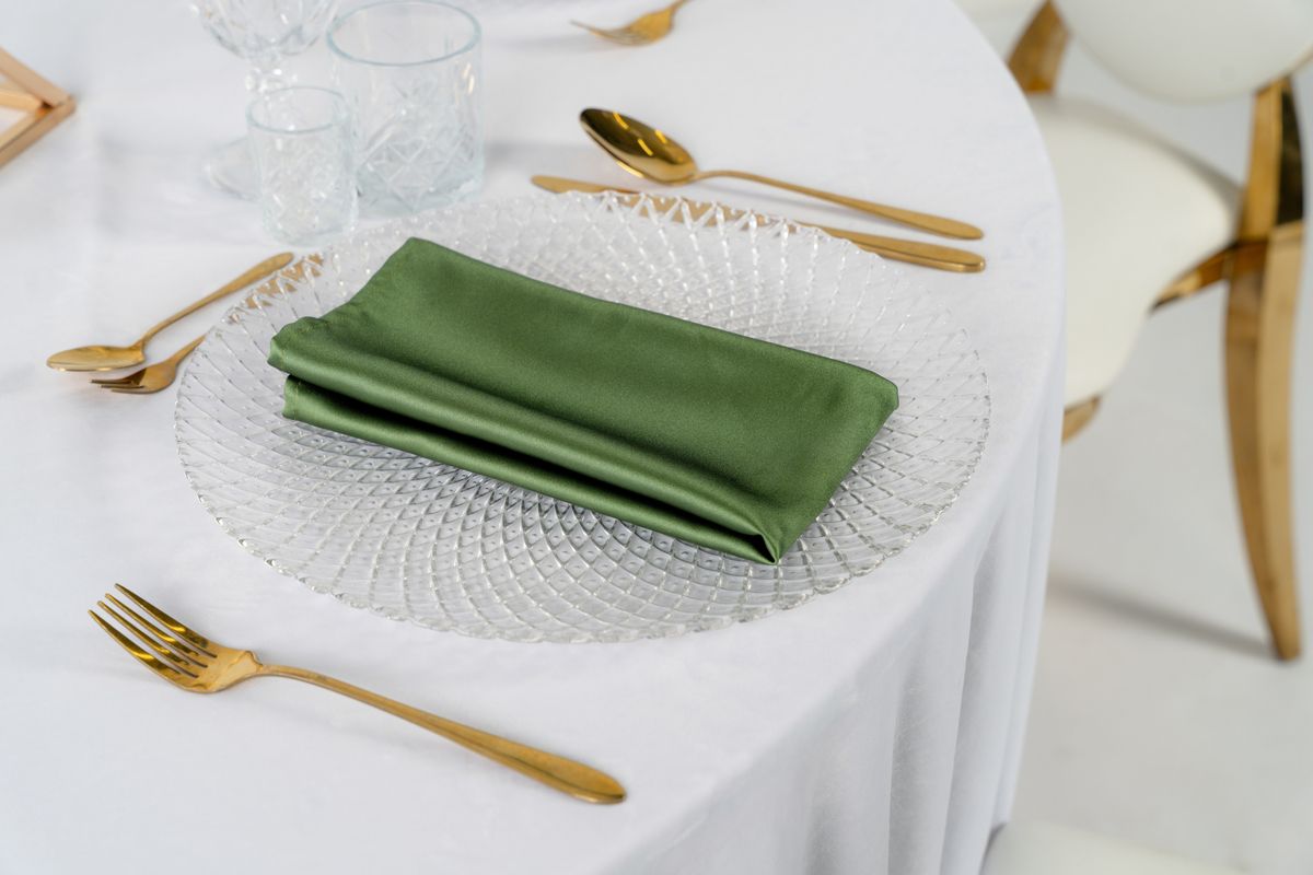 Oliwkowe serwetki satynowe na wynajem, ułożone na beżowym obrusie obok gałązek oliwnych i złotych sztućców – elegancka dekoracja stołu w stylu Tuscan Wedding